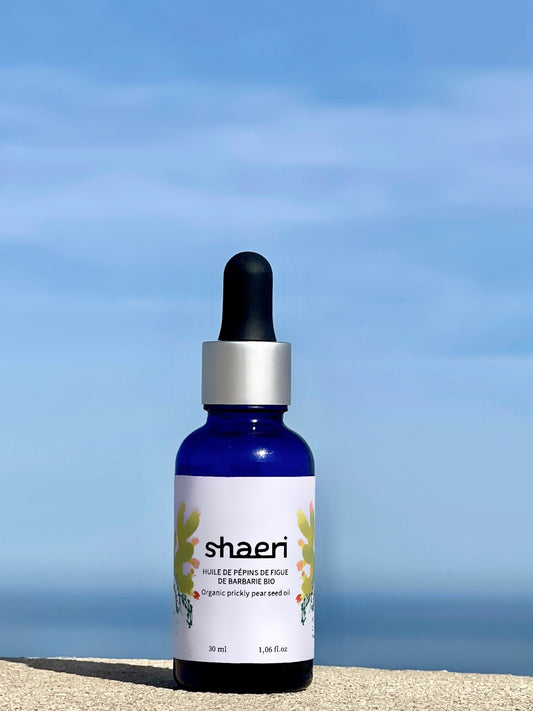 Une bouteille d'huile de pépins de figue de barbarie bio Shaeri, avec un flacon en verre bleu et un compte-gouttes noir, posée devant un ciel dégagé et une vue sur la mer.